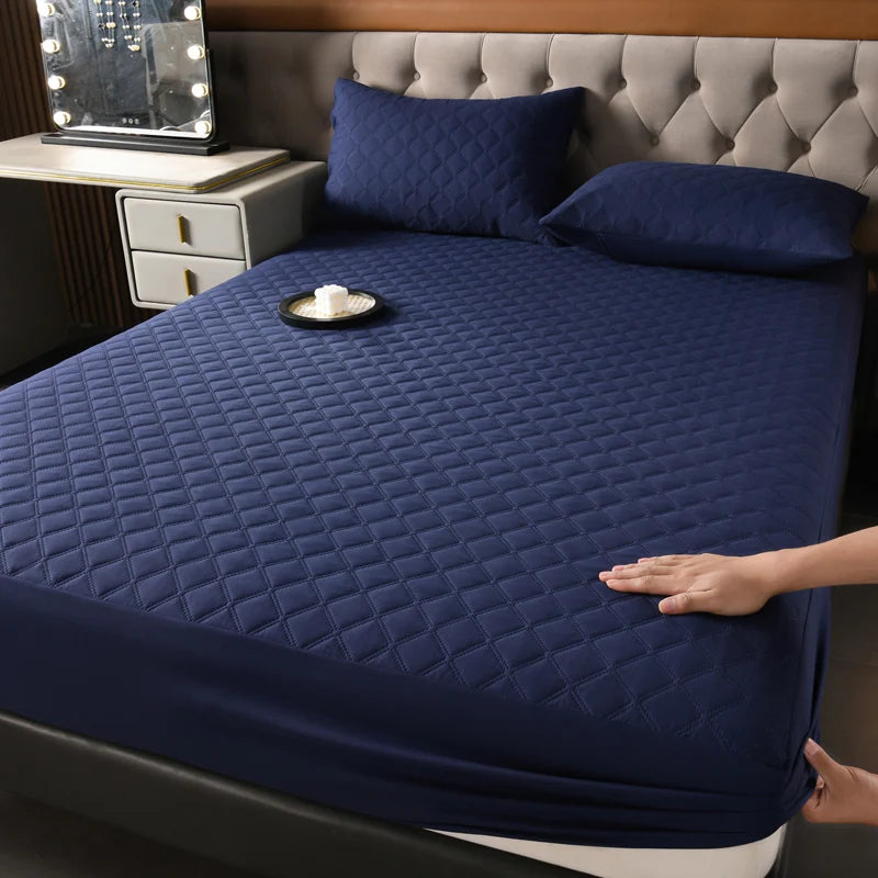 A dark blue quilted mattress protector is shown on a bed, with pillows and a bedside lamp visible.