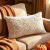 A fluffy cream throw pillow resting on a beige sofa next to an orange knitted blanket.