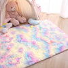 A fluffy rainbow colored plush area rug with colorful pom-poms and a teddy bear in a light pink room.