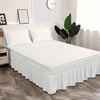A white bed with a ruffled skirt and white pillows is displayed in a bright room.