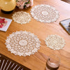 Four round, cream-colored floral embroidered lace placemats are displayed on a wooden table with a pink floral arrangement an
