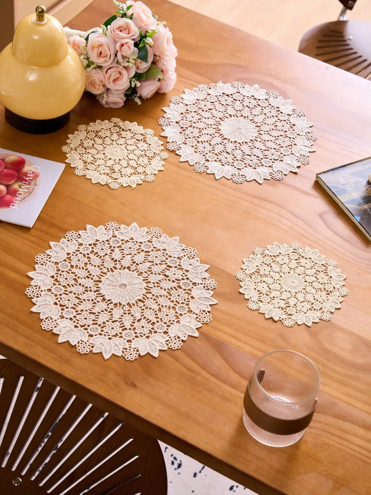 Four round, cream-colored floral embroidered lace placemats are displayed on a wooden table with a pink floral arrangement an