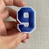 A hand holds a dark blue embroidered number 9 patch with white stitching.
