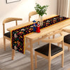 A wooden dining table set with a colorful Day of the Dead sugar skull linen table runner featuring figures and floral pattern