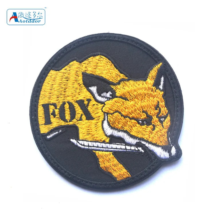 A black embroidered patch featuring a stylized golden fox head with the text FOX in yellow.
