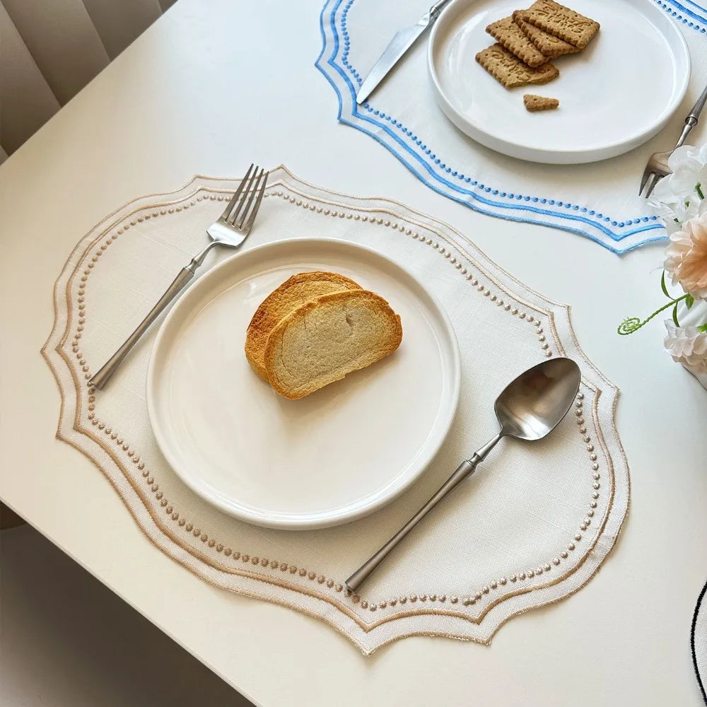A white plate with a slice of bread and silverware is centered on an embroidered linen placemat on a table.