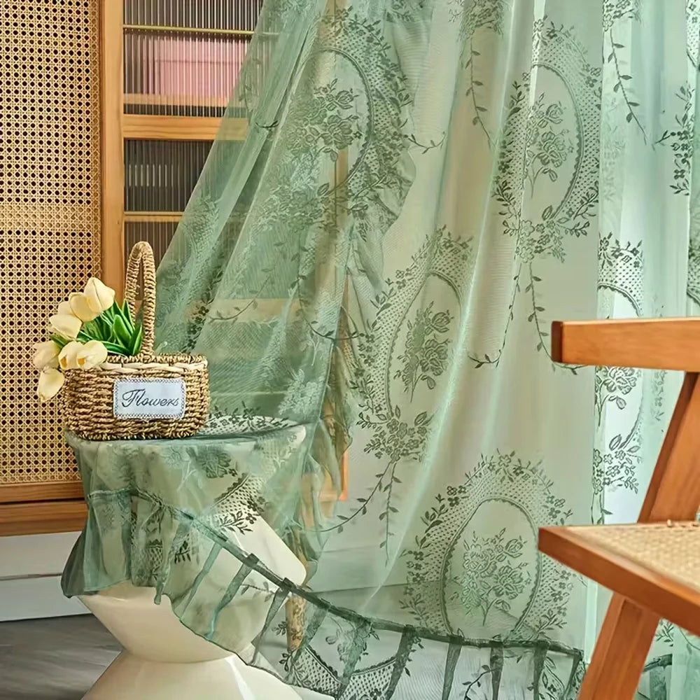 A sheer green lace floral window curtain draped over a white pedestal table.