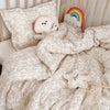 A soft, cream-colored floral print muslin blanket with a stuffed bear and a rainbow decoration on a wooden bed.