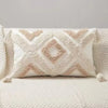 A square plush pillow cover in cream with geometric embroidery and tassel details resting on a white knitted surface.
