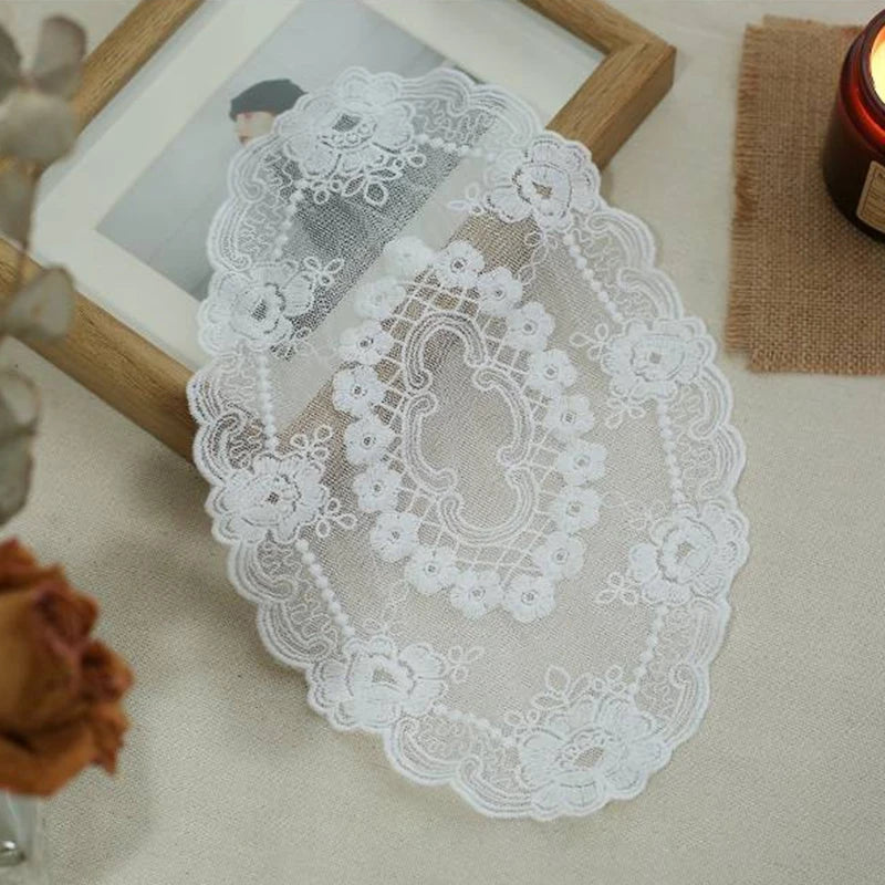 A white oval lace doily or coaster with intricate floral embroidery displayed on a light surface.