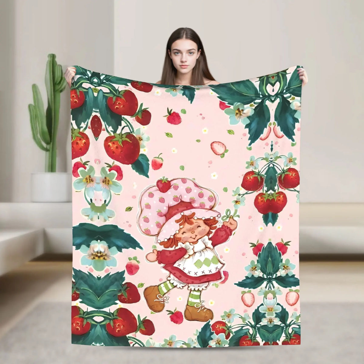 A woman holds up a pink strawberry shortcake plush throw blanket featuring strawberries and green foliage.