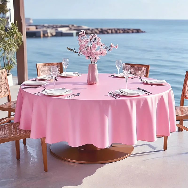 A round pink satin table overlay cloth set for an outdoor dining scene with a sea view.