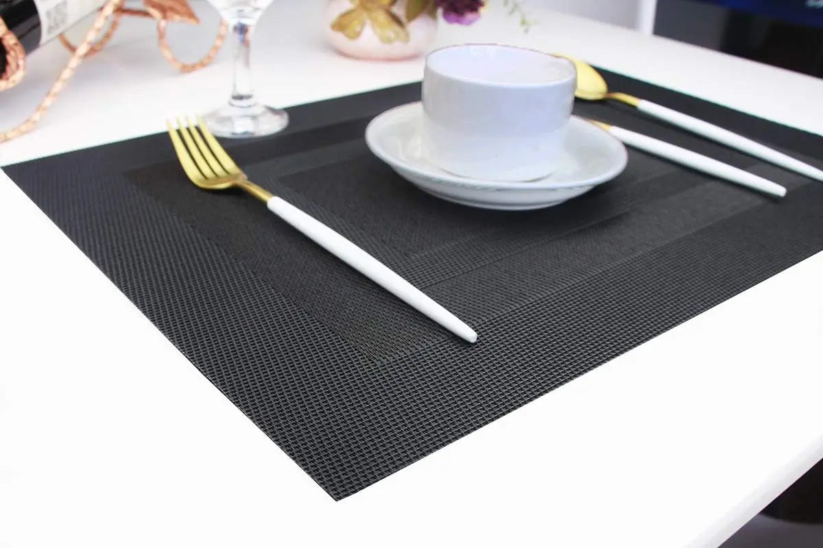 A set of six black non-slip heat resistant placemats is displayed on a white table with silverware and a white cup.