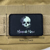 Black embroidered badge patch featuring a white skull and the text Memento Mori on a tan fabric background.