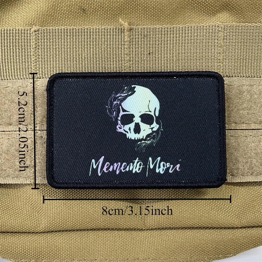 Black embroidered badge patch featuring a white skull and the text Memento Mori on a tan fabric background.