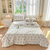 A light gray pastoral print quilt blanket is displayed on a bed with white bedding and decorative pillows.