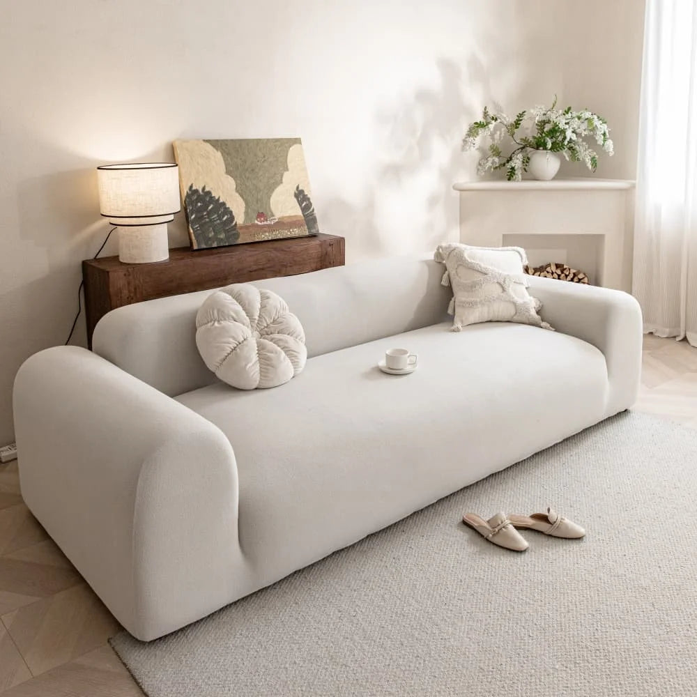 A white quilted sofa cover with elastic straps is displayed on a light-colored rug in a bright room.