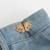 A delicate gold-colored butterfly button is attached to the denim fabric of a pant or skirt.
