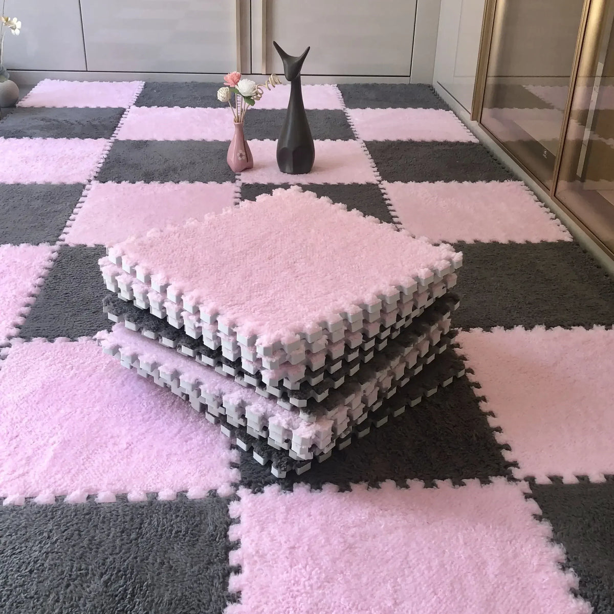 A pink and gray checkered rug mat with stacked pink fluffy mats and a black figurine on a floor.