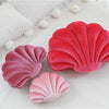 A set of three velvet stuffed sofa cushions shaped like decorative seashells in shades of pink and red resting on a white sur