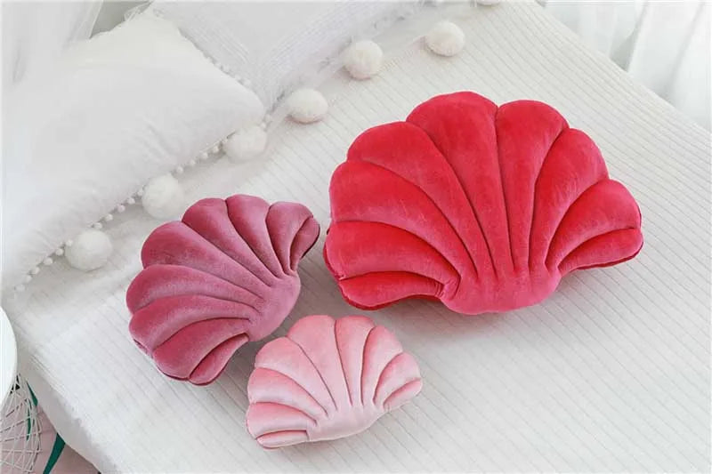 A set of three velvet stuffed sofa cushions shaped like decorative seashells in shades of pink and red resting on a white sur