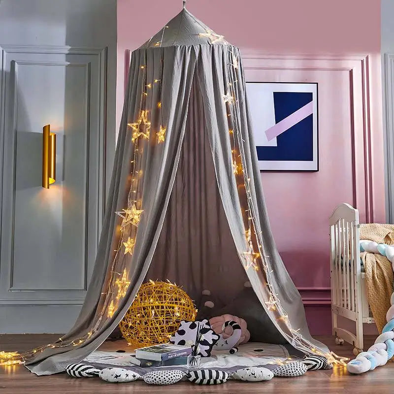 A gray princess hanging bed canopy with gold string lights and pillows in a pink room.
