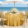 A round table set for an outdoor event with a golden tablecloth, white flowers, and gold chairs by the sea.