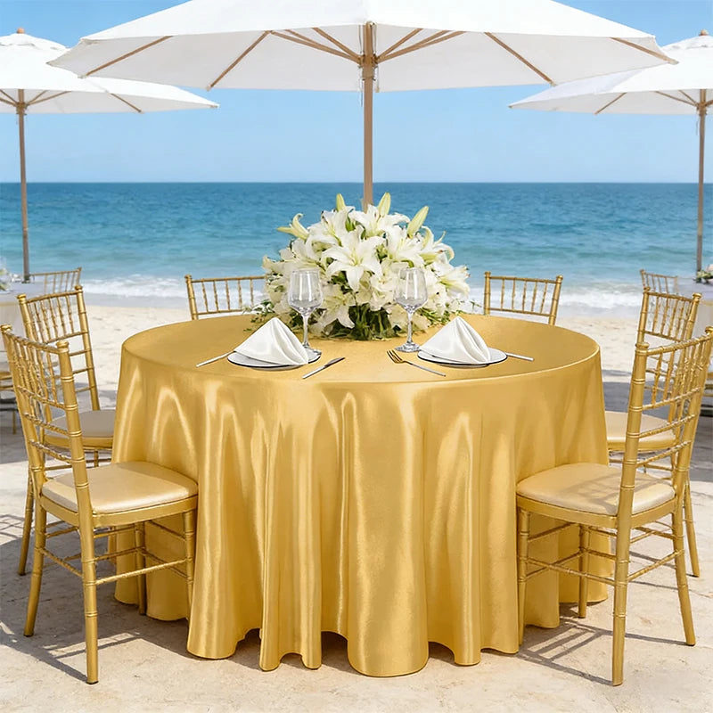 A round table set for an outdoor event with a golden tablecloth, white flowers, and gold chairs by the sea.