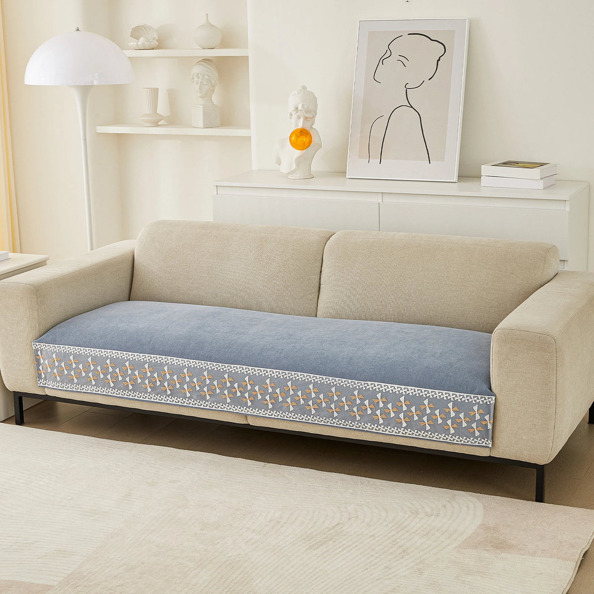 A beige sofa with a blue quilted cover featuring a white lace trim, displayed in a bright living room.