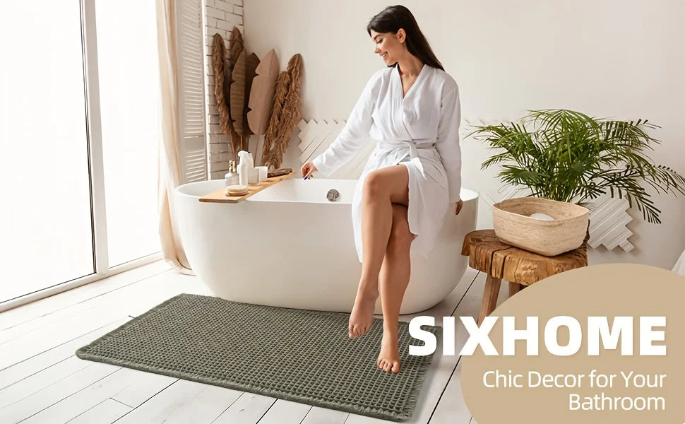 A woman sits in a white bathtub surrounded by natural decor, with a green waffle bath mat on the floor.