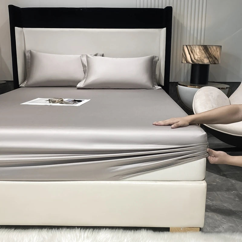 A luxury bed with a fitted sheet and pillows in silver-gray tones, featuring a dark headboard and modern decor.
