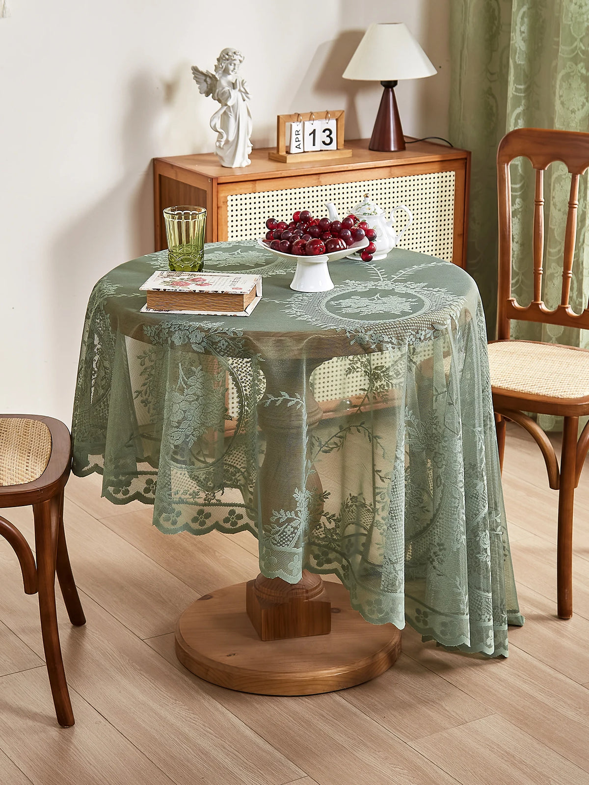 A round table set with a green floral lace tablecloth, a white lamp, and a bowl of red berries.