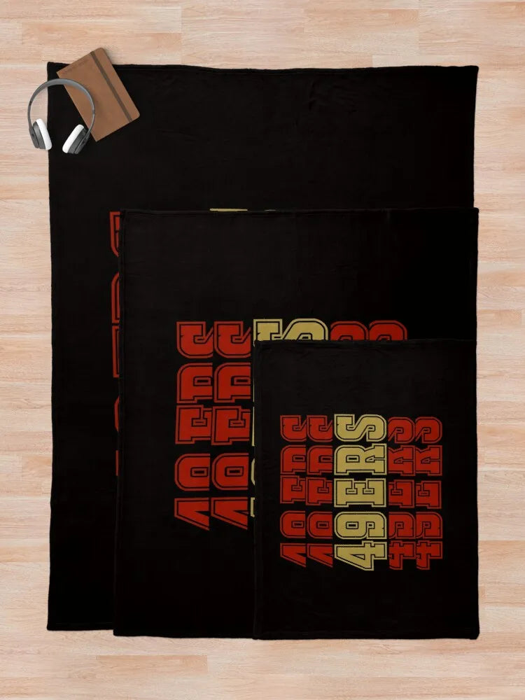 A black knitted throw blanket with colorful lettering is displayed on a light wooden surface.