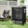 A black waterproof bird cage with a mesh night cover is placed on a green lawn next to a wooden bench and white fence.