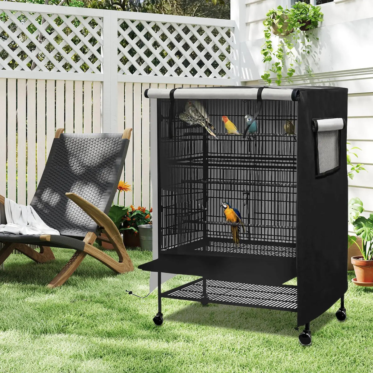 A black waterproof bird cage with a mesh night cover is placed on a green lawn next to a wooden bench and white fence.