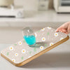 A hand places a glass of blue liquid onto a wooden silicone drawer mat with a white and green floral pattern.