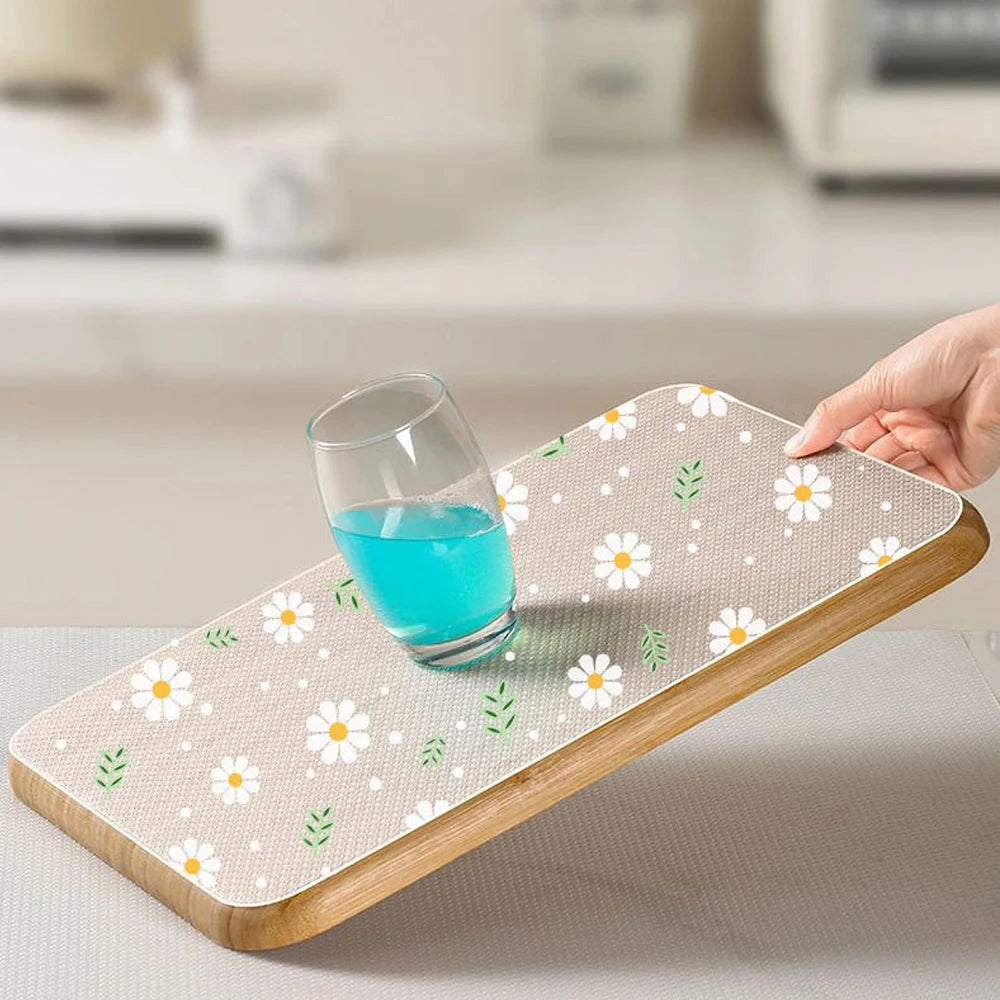 A hand places a glass of blue liquid onto a wooden silicone drawer mat with a white and green floral pattern.