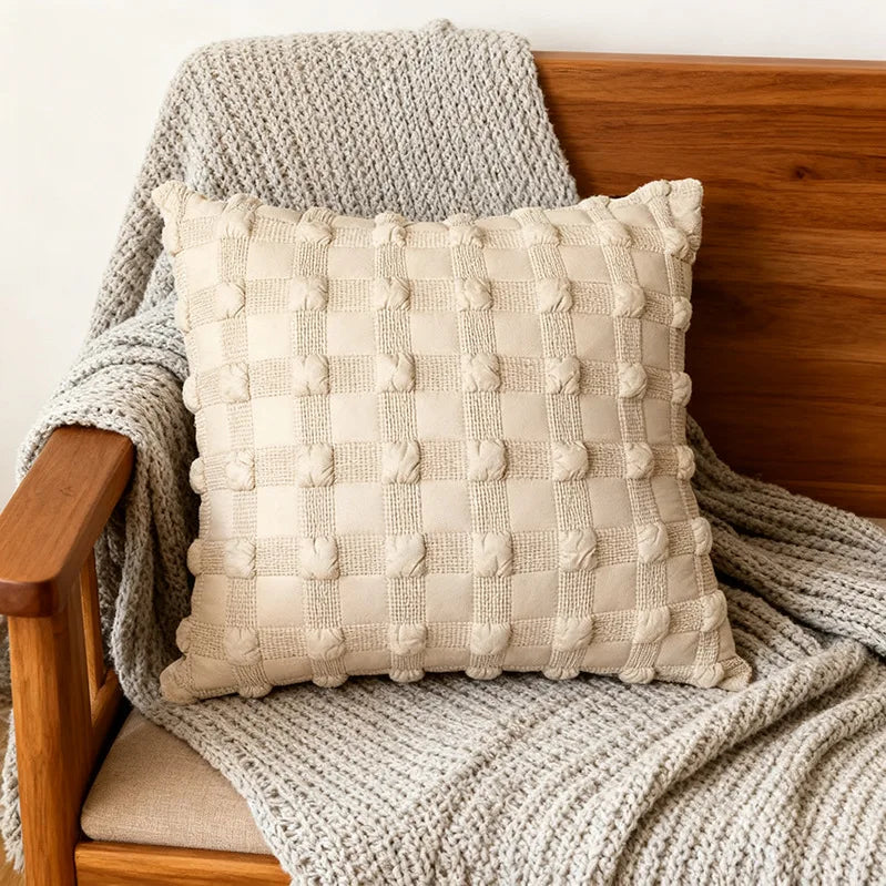 A square beige quilted pillow with textured stitching resting on a wooden surface covered by a gray knit blanket.
