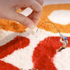 A hand is interacting with an irregular shaped plush floor rug featuring orange, white, and red patterns.