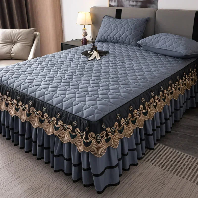 A quilted bed skirt in gray with gold embroidery and pleats, shown on a bed.