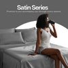 A woman is sitting on a bed with white satin bedding, holding a dark object to her ear.
