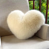 A fluffy, heart-shaped throw pillow made of light beige fox fur resting on a sofa near a window.
