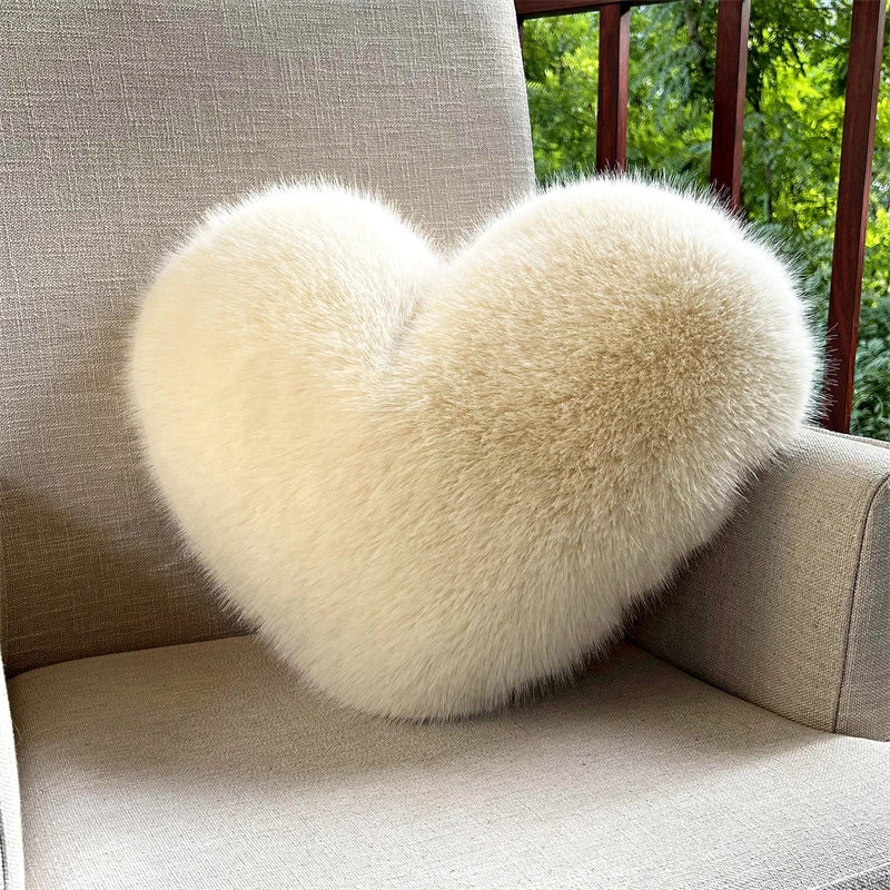 A fluffy, heart-shaped throw pillow made of light beige fox fur resting on a sofa near a window.