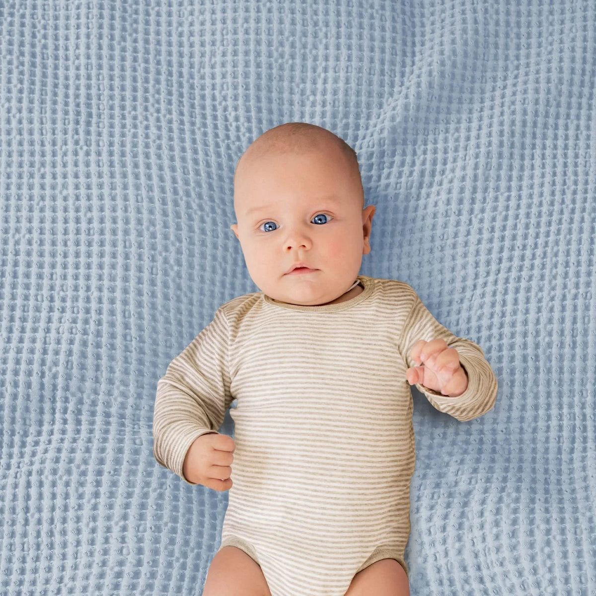 A baby wearing a beige striped onesie lying on a light blue waffle blanket.