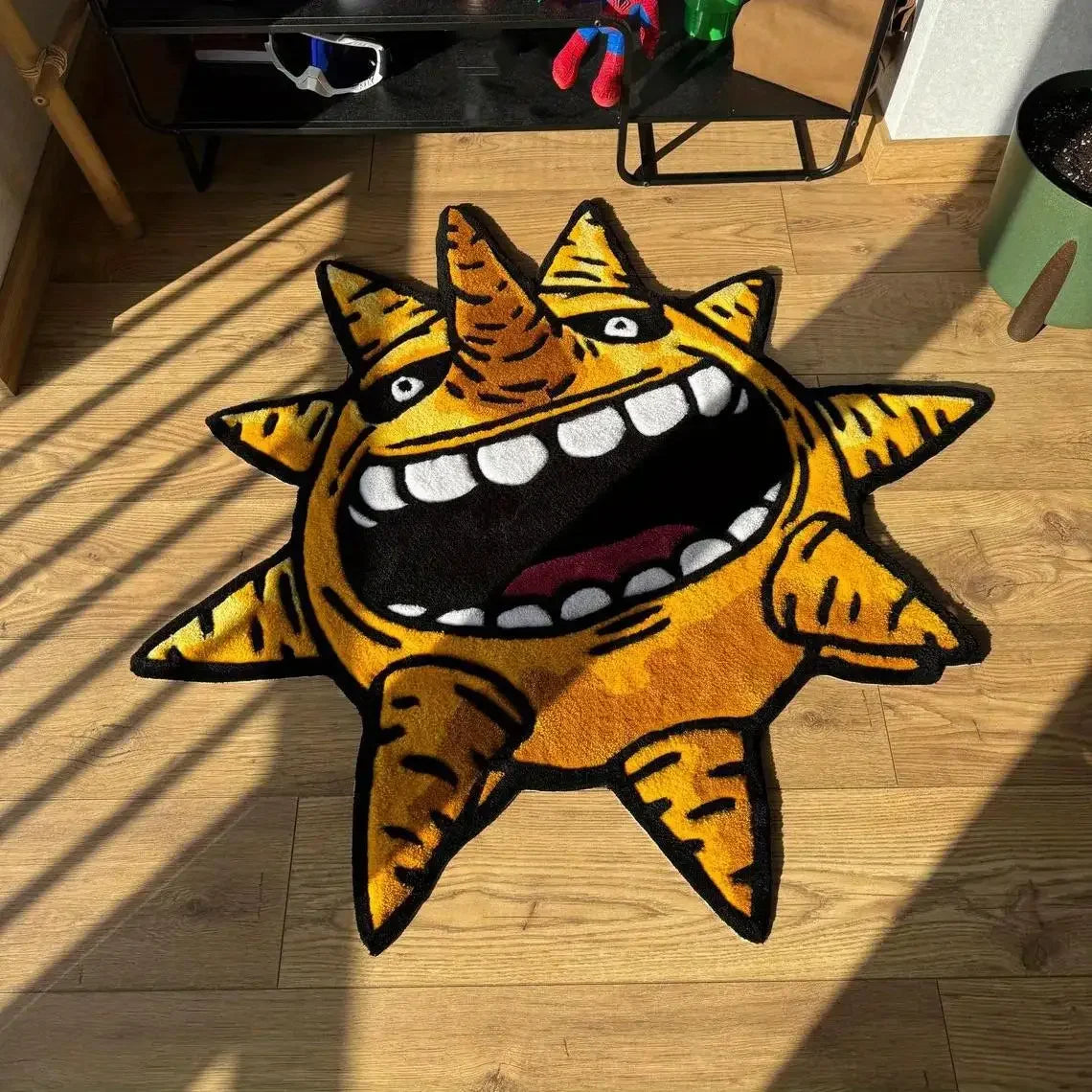 A yellow and orange sun motif floor mat with a fierce expression is placed on a wooden floor.