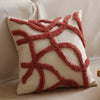 A square cream colored plush pillow cover with geometric lines embroidered in a reddish-brown tassel detail.