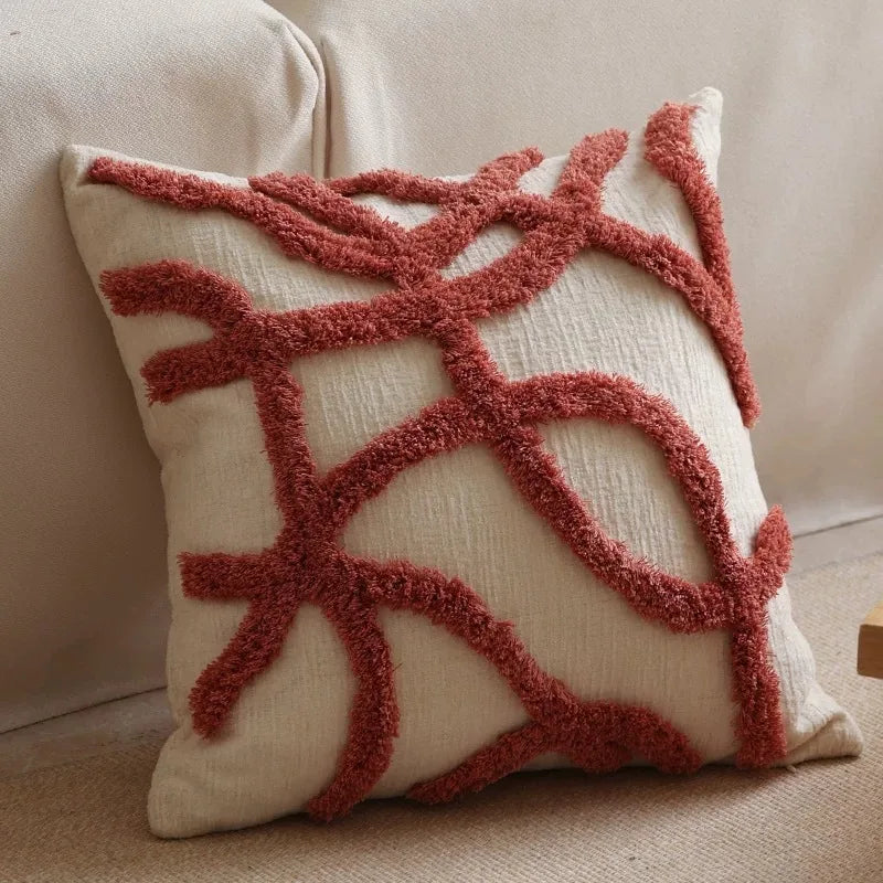 A square cream colored plush pillow cover with geometric lines embroidered in a reddish-brown tassel detail.