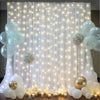 A sheer white tulle backdrop decorated with warm string lights, surrounded by clear and white balloons and floral arrangement