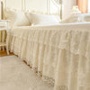 A white lace ruffled bed skirt set draped over a bed with light-colored bedding and a decorative headboard.