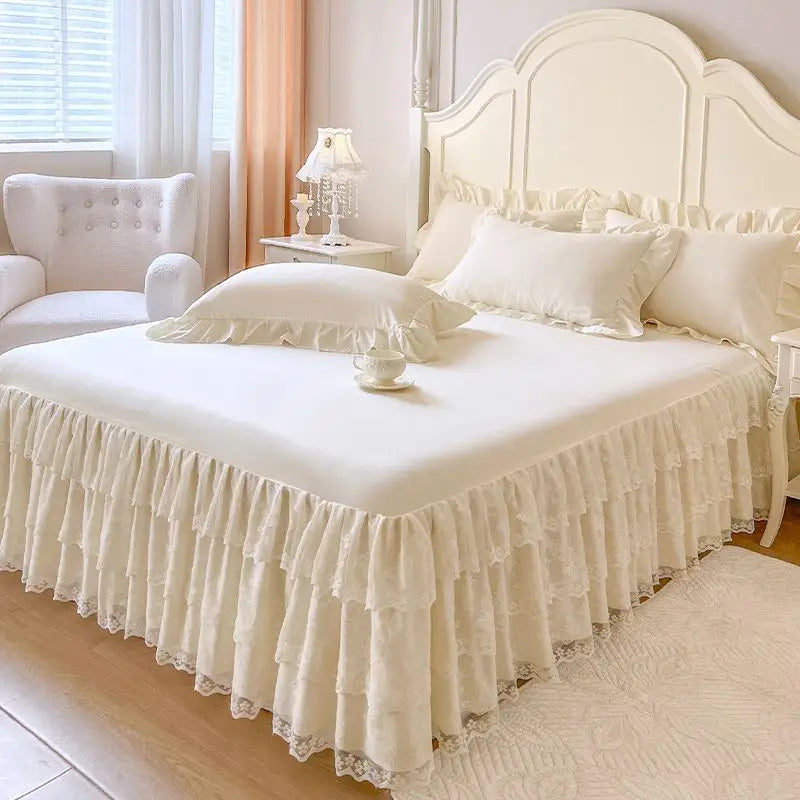 A white bed with a lace ruffled skirt set featuring a tall upholstered headboard and white linens.
